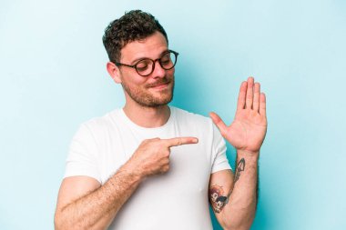 Mavi arka planda izole edilmiş genç beyaz adam gülümseyerek beş numarayı parmaklarıyla gösteriyor..