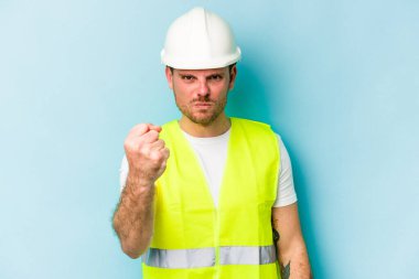 Mavi arka planda izole edilmiş genç işçi kameraya yumruk, agresif yüz ifadesi gösteriyor..