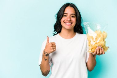 Mavi arka planda bir paket cipsi tutan genç İspanyol kadın gülümsüyor ve başparmağını kaldırıyor.