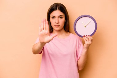 Latin kökenli genç bir kadın bej arka planda izole edilmiş bir saat tutuyor elini uzatmış, dur işaretini gösteriyor ve seni engelliyor..