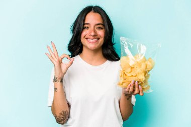 Genç İspanyol kadın elinde bir paket cipsle mavi arka planda izole edilmiş neşeli ve kendine güveni tam bir jest yapıyor..