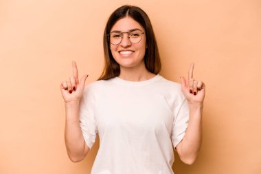 Latin kökenli genç bir kadın bej arka planda izole edilmiş. İki parmağı da boş bir alan gösteriyor..