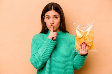 Genç İspanyol kadın elinde bir çanta dolusu cipsle bej arka planda izole edilmiş bir şekilde bir sır saklıyor ya da sessizlik istiyor..