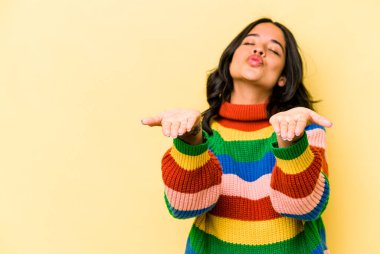 Sarı arka planda izole edilmiş genç İspanyol kadın dudakları katlıyor ve hava öpücüğü göndermek için avuç içleri tutuyor..