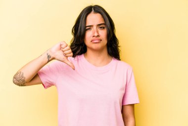 Sarı arka planda izole edilmiş genç İspanyol kadın baş parmağını aşağı gösteriyor, hayal kırıklığı konsepti..