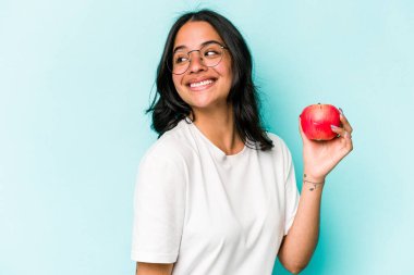 Mavi arka planda izole edilmiş bir elma tutan genç İspanyol kadın gülümseyen, neşeli ve keyifli bir kenara bakıyor..