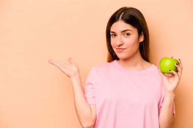 Elinde bej arka planda izole edilmiş bir elma tutan genç İspanyol kadın bir avuç içinde fotokopi alanı ve belinde başka bir el tutuyor..