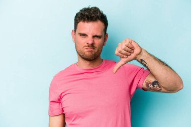 Mavi arka planda izole edilmiş genç beyaz bir adam baş parmağını aşağı gösteriyor ve hoşlanmadığını ifade ediyor..