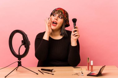 Makyaj yapan genç bir kadın pembe arka planda izole edilmiş bir makyaj dersi veriyor. Bağırıyor ve avuç içini açık tutuyor..