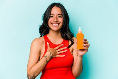 Mavi arka planda güneş kremi tutan genç İspanyol kadın elini göğsünde tutarak kahkahalar atıyor..