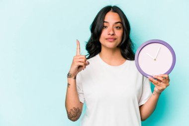 Mavi arka planda bir saat tutan genç İspanyol kadın bir numarayı parmağıyla gösteriyor..