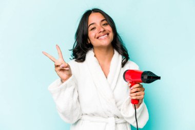Mavi arka planda izole edilmiş kurutma makinesini tutan genç İspanyol kadın neşeli ve kaygısız parmaklarıyla barış sembolü gösteriyor..