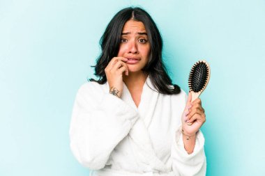 Mavi arka planda saç fırçası tutan genç İspanyol kadın tırnaklarını yiyor, gergin ve çok endişeli..
