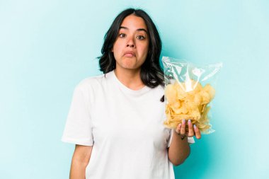 Mavi arka planda izole edilmiş bir paket cipsi tutan genç İspanyol kadın omuzlarını silker ve gözleri açık kalır..