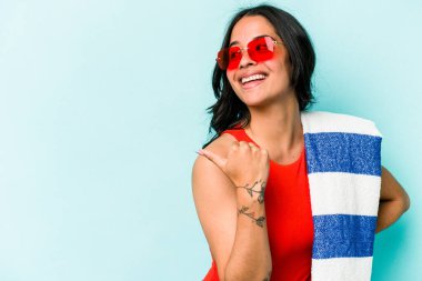 Plaj havlusunu tutan genç İspanyol kadın mavi arka plan noktalarında izole edilmiş başparmak uzakta, gülüyor ve kaygısız.