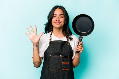 Genç İspanyol tencereli kadın elinde tavayla mavi arka planda izole edilmiş gülümseyen beş numarayı parmaklarıyla gösteriyor..