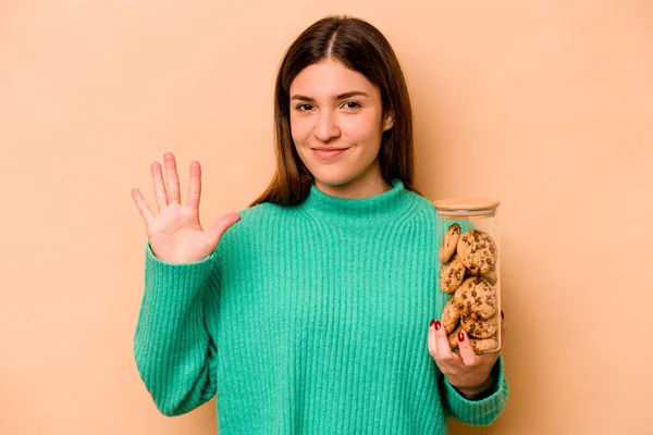 Elinde kurabiye kavanozu tutan genç İspanyol kadın bej arka planda izole edilmiş gülümseyen beş numarayı parmaklarıyla gösteriyor..