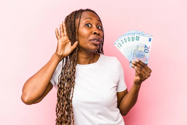 Young African American woman holding banknotes isolated on pink background trying to listening a gossip.
