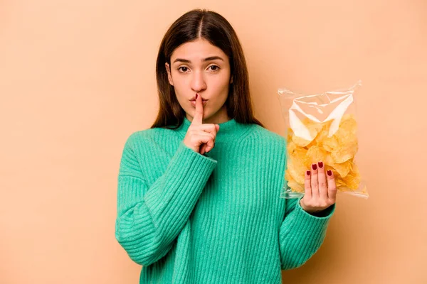 Genç İspanyol kadın elinde bir çanta dolusu cipsle bej arka planda izole edilmiş bir şekilde bir sır saklıyor ya da sessizlik istiyor..