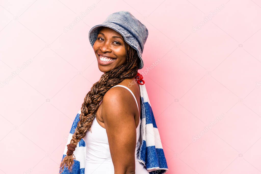Young African American woman holding a towel isolated on pink background looks aside smiling, cheerful and pleasant.