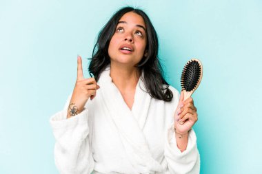 Mavi arka planda saç fırçası tutan genç İspanyol kadın ağzı açık bir şekilde üst tarafı işaret ediyor..