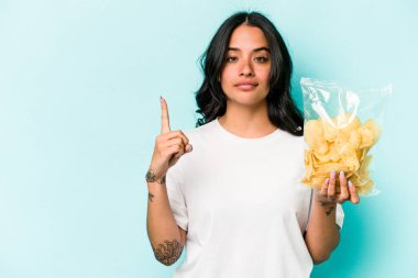 Mavi arka planda bir çanta dolusu cipsi tutan genç İspanyol kadın bir numarayı parmağıyla gösteriyor..