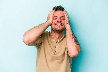 Mavi arka planda izole edilmiş genç beyaz adam ellerini başının üstünde tutarak neşe içinde gülüyor. Mutluluk kavramı.