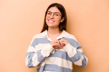 Latin kökenli genç bir kadın bej arka planda izole edilmiş gülümsüyor ve elleriyle kalp şekli gösteriyor..