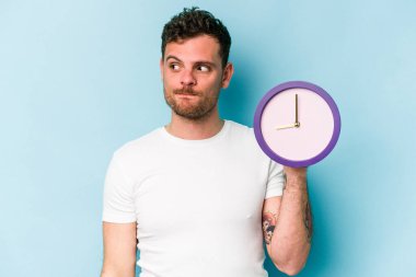 Kafkasyalı genç bir adam mavi arka planda izole edilmiş bir saat tutuyor. Kafası karışmış ve kendinden emin değil..