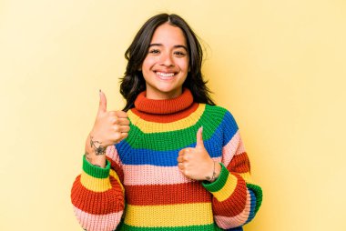 Sarı arka planda izole edilmiş genç İspanyol kadın iki parmağını da kaldırıyor, gülümsüyor ve kendinden emin..