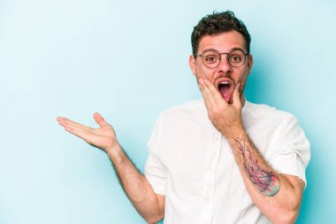 Mavi arka planda izole edilmiş genç beyaz bir adam avucunun içinde fotokopi alanı tutuyor, el üstünde tutuyor. Şaşırmış ve mutlu..