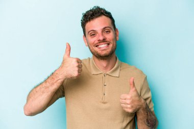 Kafkasyalı genç adam mavi arka planda izole edilmiş. Baş parmağını kaldırıyor, gülümsüyor ve kendinden emin..