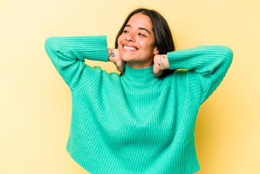 Sarı arka planda izole edilmiş genç İspanyol kadın kendini güvende hissediyor, elleri başının arkasında..