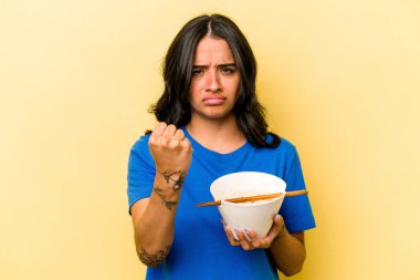 Mavi arka planda ramen yiyen genç İspanyol kadın kameraya yumruğunu gösteriyor, agresif yüz ifadesi..