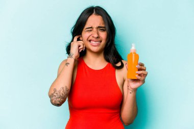 Mavi arka planda güneş kremi tutan genç İspanyol kadın kulaklarını elleriyle kapatıyor..