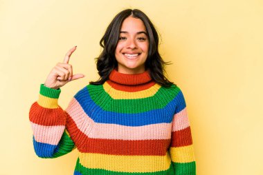 Sarı arka planda izole edilmiş genç İspanyol kadın işaret parmaklarıyla küçük bir şey tutuyor, gülümsüyor ve kendinden emin..