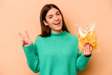 Latin kökenli genç bir kadın bej arka planda izole edilmiş bir çanta tutuyor neşeli ve kaygısız parmakları olan barış sembolü gösteriyor..