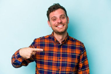 Mavi arka planda izole edilmiş genç beyaz adam bir gömlek fotokopi alanını işaret ediyor, gururlu ve kendinden emin.