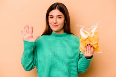 Bej arka planda izole edilmiş bir paket cipsi tutan genç İspanyol kadın gülümseyerek beş numarayı parmaklarıyla gösteriyor..