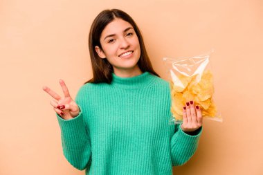 Latin kökenli genç bir kadın elinde bir çanta dolusu cipsle bej arka planda iki numarayı parmaklarıyla gösteriyor..