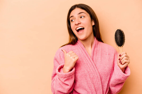 Young hispanic woman holding hairbrush isolated on beige background points with thumb finger away, laughing and carefree.