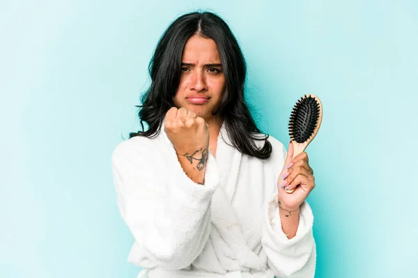 若いヒスパニック系の女性は カメラに拳を示す青い背景に隔離されたヘアブラシを保持します 積極的な顔の表情 — ストック写真