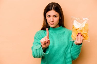 Latin kökenli genç bir kadın elinde bir çanta dolusu cipsle bej arka planda bir numarayı parmağıyla gösteriyor..