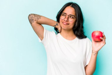 Mavi arka planda izole bir elma tutan genç İspanyol kadın kafasının arkasına dokunuyor, düşünüyor ve bir seçim yapıyor..