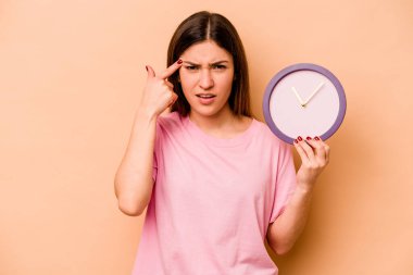 Latin kökenli genç bir kadın bej arka planda izole edilmiş bir saat tutuyor işaret parmağıyla yaptığı hayal kırıklığı jestini gösteriyor..