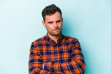 Kafkasyalı genç adam mavi arka planda izole edilmiş hoşnutsuzca kaşlarını çatmış, kollarını kavuşturmuş..