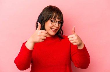 Pembe arka planda izole edilmiş genç beyaz kadın iki parmağını da kaldırıyor, gülümsüyor ve kendinden emin..