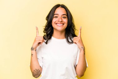 Sarı arka planda izole edilmiş genç İspanyol kadın iki parmağı da boş bir alan gösteriyor..
