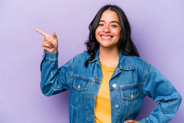 Mor arka planda izole edilmiş genç İspanyol kadın işaret parmağıyla işaret ederek gülümsüyor..