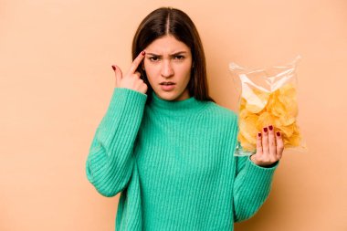 Latin kökenli genç bir kadın, elinde bej arka planda izole edilmiş bir paket cipsle işaret parmağıyla hayal kırıklığı gösteren bir jest yapıyor..